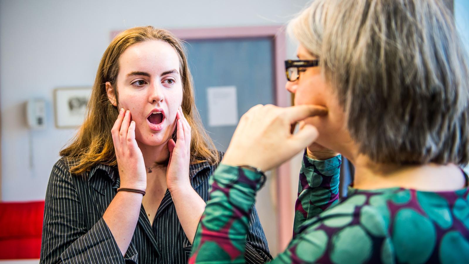 Blick in eine Unterrichtssituation, in der eine Gesangslehrerin einer Studentin die ideale Mundöffnung beim Singen vorzeigt.