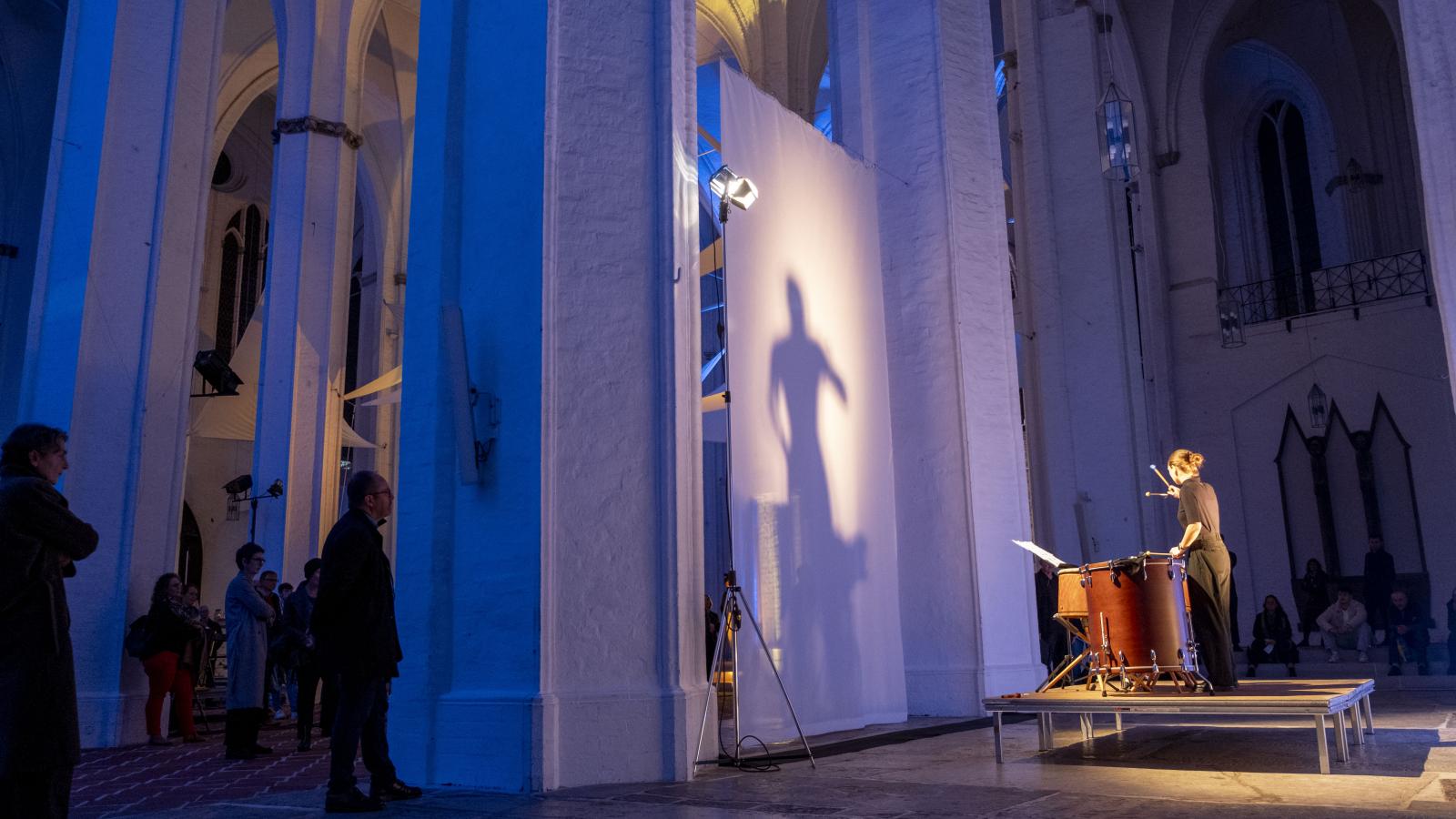 Blick auf einen Schlagzeuger, der in einer blau beleuchteten Kirche vor einer Leinwand spielt, die seinen Schatten reflektiert