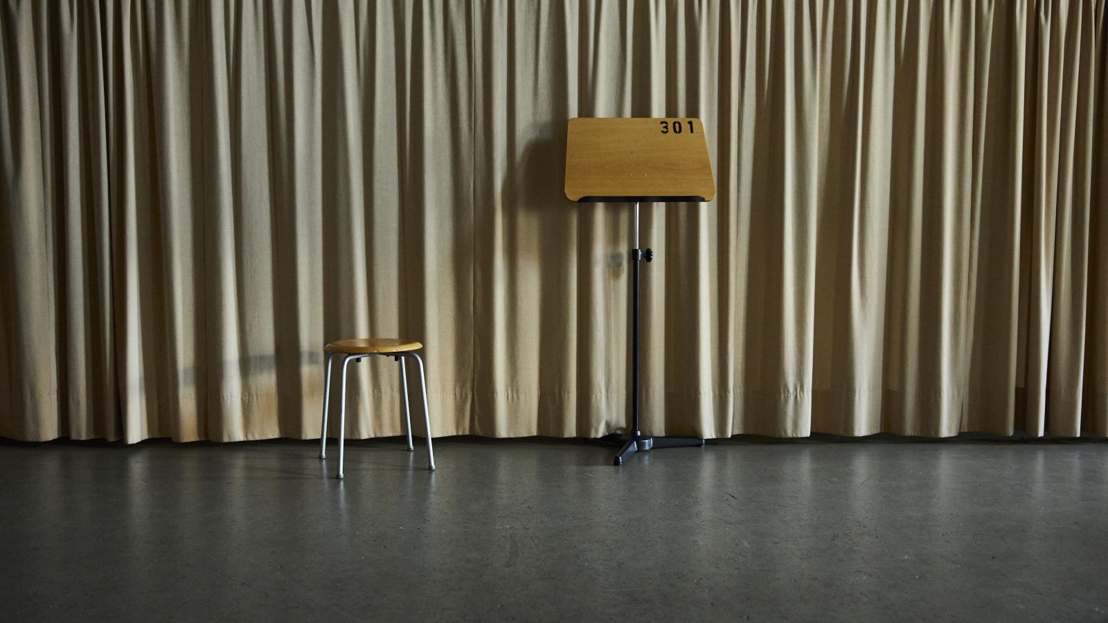 Blick auf einen Hocker neben einem Notenständer vor einem langen, hellen Vorhang