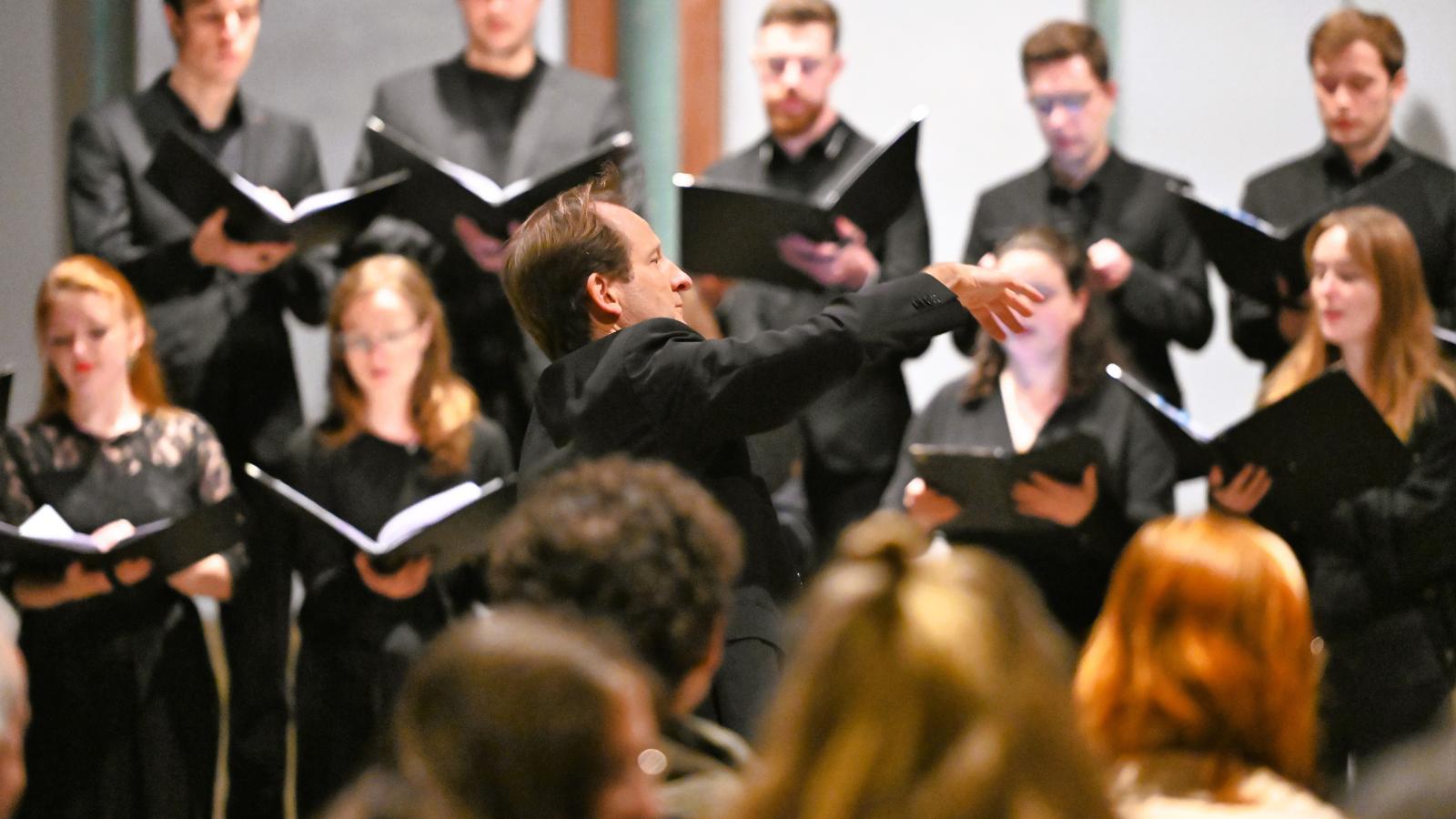 Johannes Knecht dirigiert, umgeben von Sängerinnen und Sängern, schwarze Konzertkleidung, Publikum im Vordergrund
