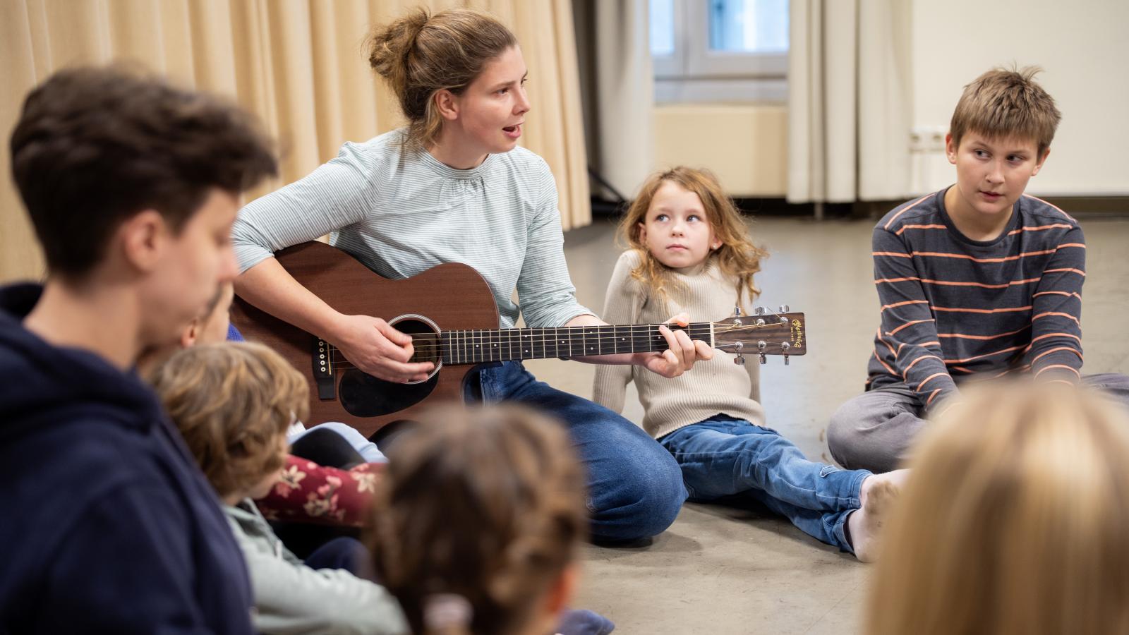 Blick auf eine Studentin, die an der Gitarre Musik mit Grundschülerinnen und -schülern macht
