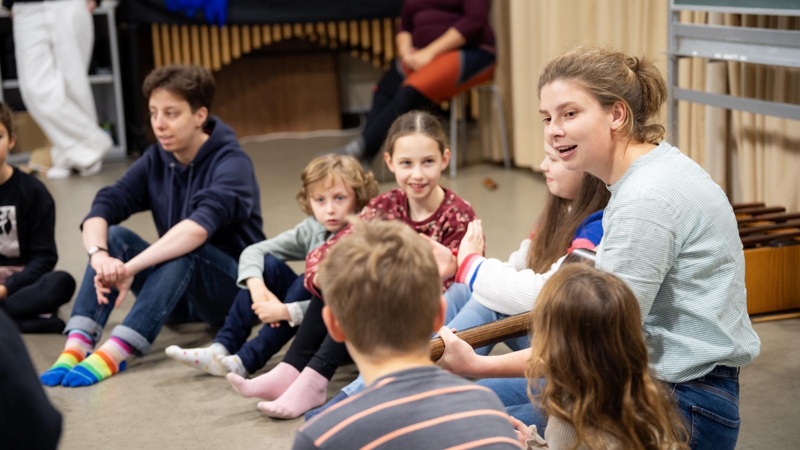 Blick auf eine Gruppe von Schülerinnen und Schülern, die mit einer Studentin zusammen Musik machen.