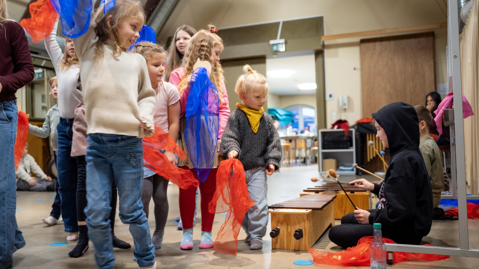 Blick auf eine Gruppe von Schülerinnen und Schülern, die mit einer Studentin zusammen Musik machen.