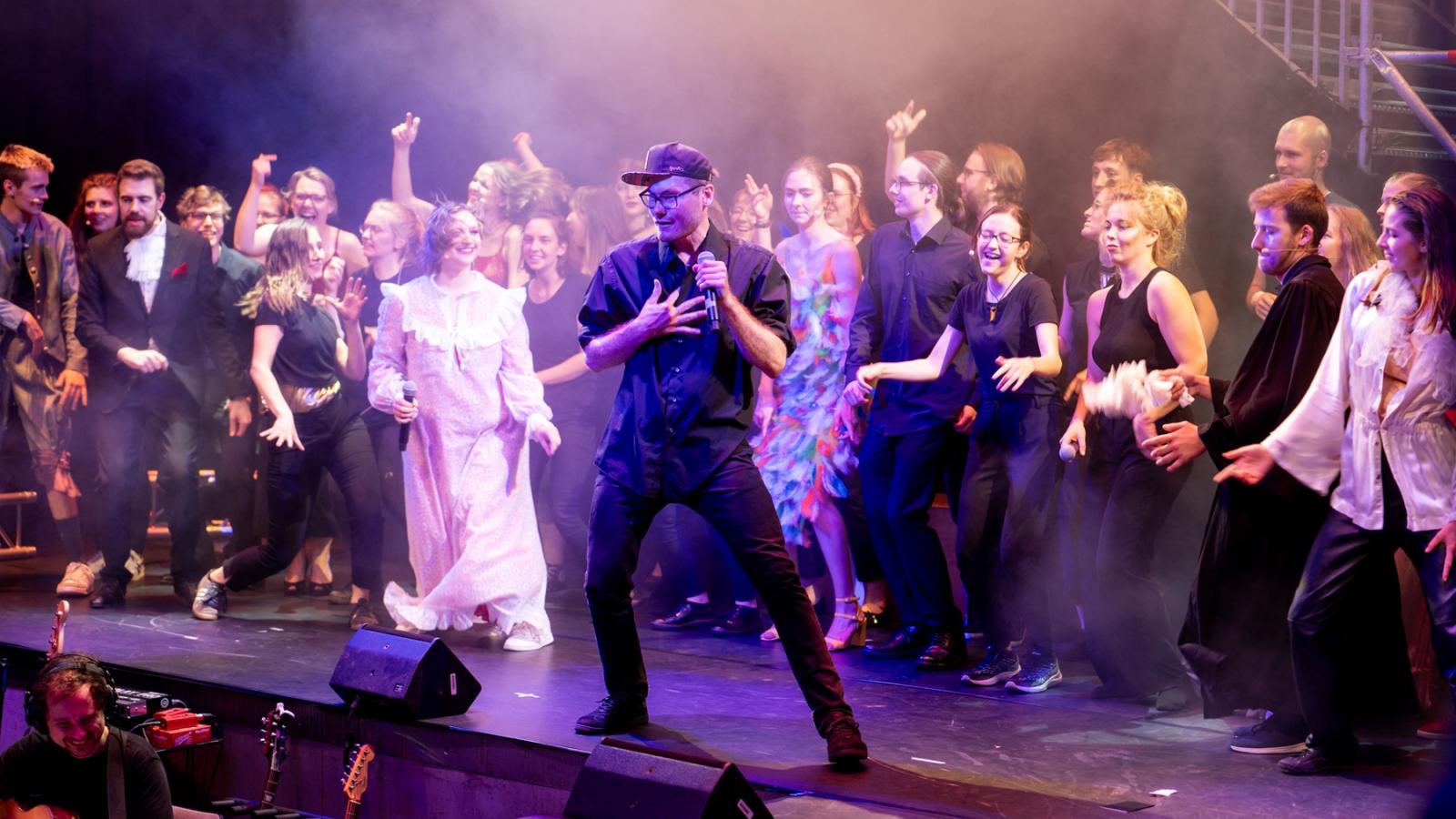 Studierende auf der Bühne in einem halbkreis angeordnet, alle singend und in Bewegung, in der Mitte des halbkreises und so auch im vordergrund ein student mit mikro in der hand in ganz schwarz. 