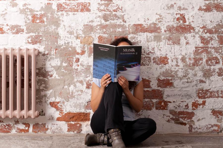 Eine Person sitzt auf einem Gang an eine alte Flurmauer gelegt und hält ein Buch vor das Gesicht.