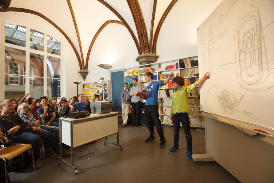 Jugendliche stehen in einem MUsikraum vor einem Schaubild und erklären ihren Mitschüler:innen, wie eine Tuba funktioniert.