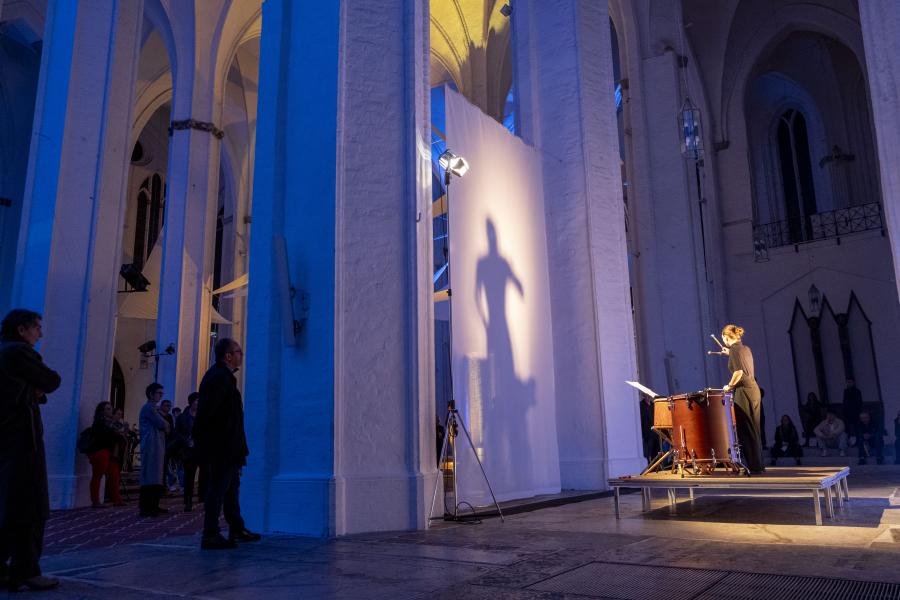 Blick auf einen Schlagzeuger, der in einer blau beleuchteten Kirche vor einer Leinwand spielt, die seinen Schatten reflektiert