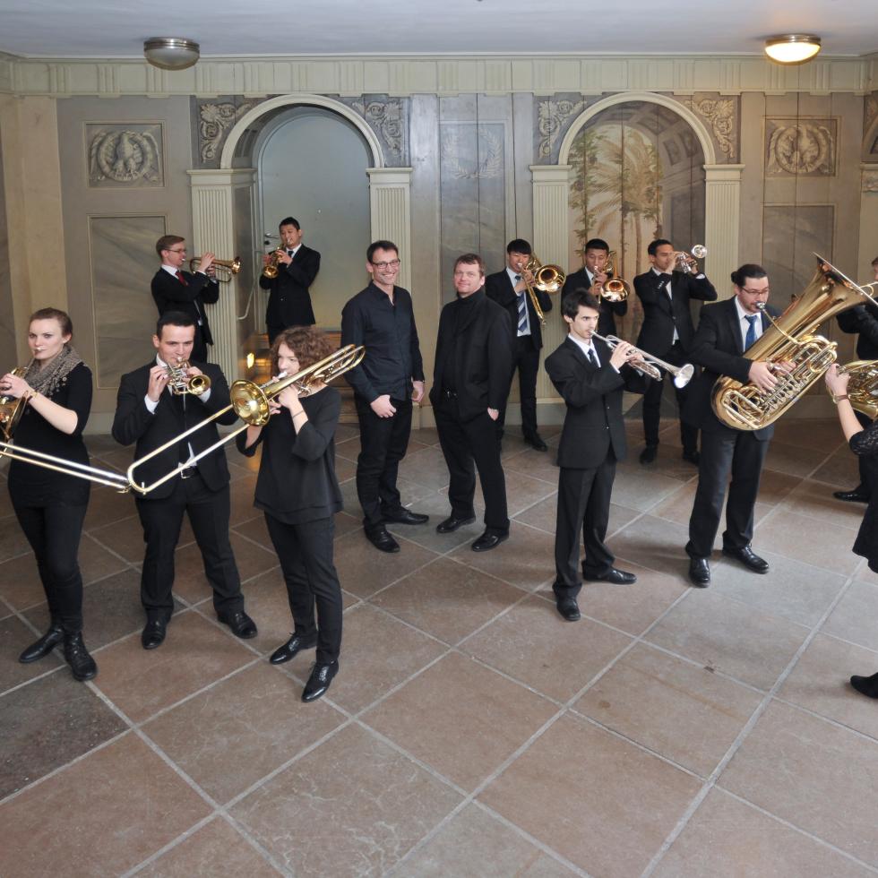 Studierende stehen in der Empirediele der MHL in einer losen Gruppe, auf ihren Instrumenten blasend, die Dozierenden in der Mitte